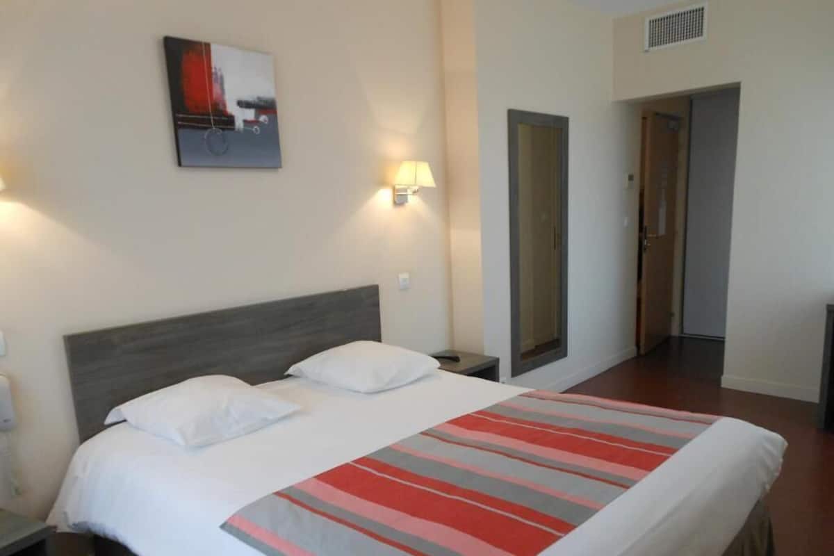 A hotel room with a bed, a nightstand, a lamp, a painting on the wall, and a door leading to another room.