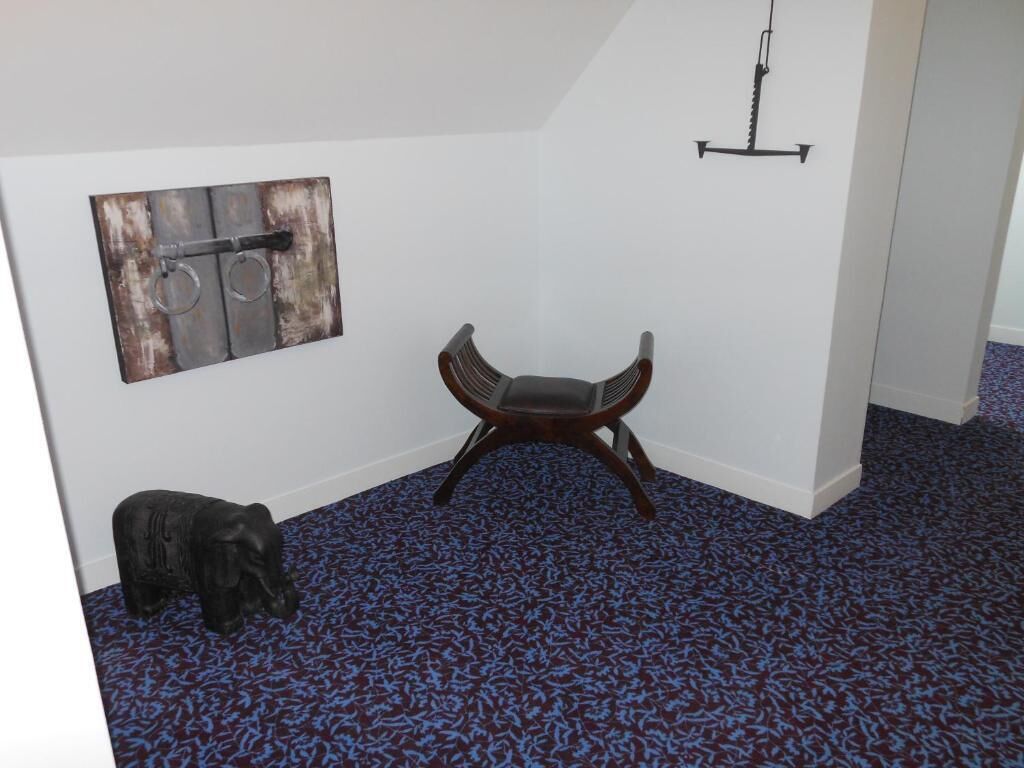 A room with a blue patterned carpet, a wooden chair, a wall-mounted artwork of a metal object, and a small elephant statue.