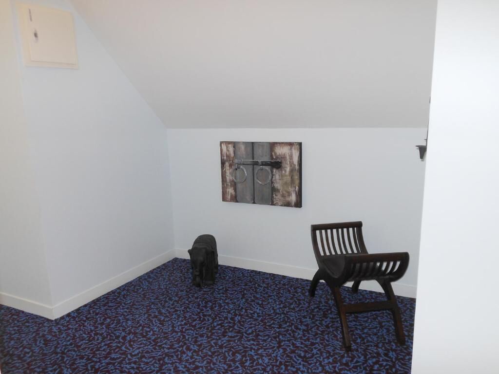 Une pièce avec un plafond en pente, une moquette bleue à motifs, une chaise noire et une œuvre d’art rustique accrochée au mur.