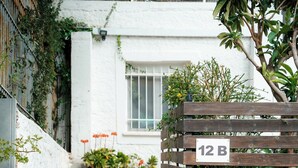 Un mur de pierres avec une porte en bois, un bâtiment blanc doté d’une fenêtre et une clôture en bois sur laquelle est affiché un panneau indiquant « 12 B ».