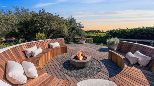Eine runde Holzterrasse mit einem Feuerkorb, Sitzbereichen und Kübelpflanzen.