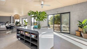 Ein moderner Wohnraum mit einer zentralen Kücheninsel, Bücherregalen, einem Deckenventilator und großen Fenstern.