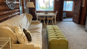Et hyggeligt interiør i en træhytte med en beige sofa, en grøn skammel, et spisebord med stole og en loftsvifte.
