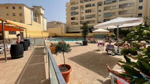 A rooftop pool area with lounge chairs, potted plants, and a building in the background.