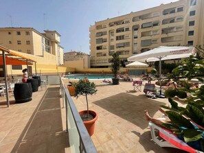 Uma área de piscina no terraço com espreguiçadeiras, plantas em vasos e um edifício ao fundo.