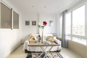 A modern hotel room with a bed, a small table, a vase with flowers, and a large window.