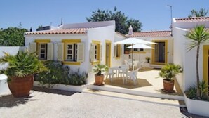Ein weißes Gebäude mit gelben Akzenten, eine Terrassenfläche mit weißen Stühlen und einem Tisch sowie Kübelpflanzen.