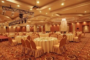 Un amplio salón de banquetes con mesas redondas, sillas y un escenario con equipo de iluminación.