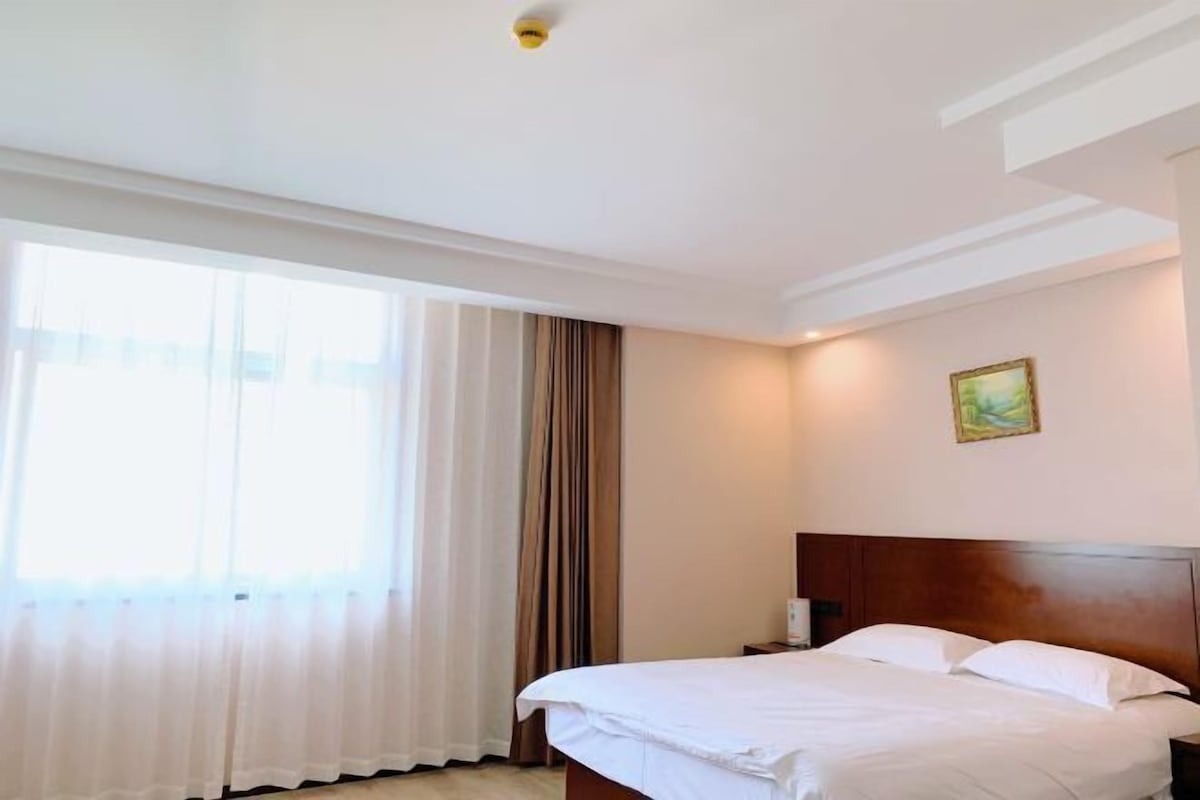 A hotel room with a bed, a nightstand, a painting on the wall, and a window with curtains.