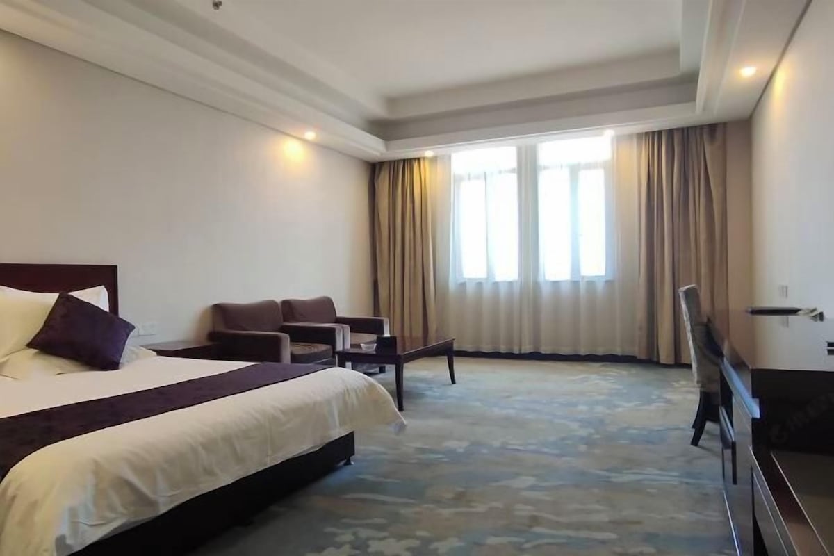 A hotel room with a large bed, two armchairs, a small table, and a window with curtains.