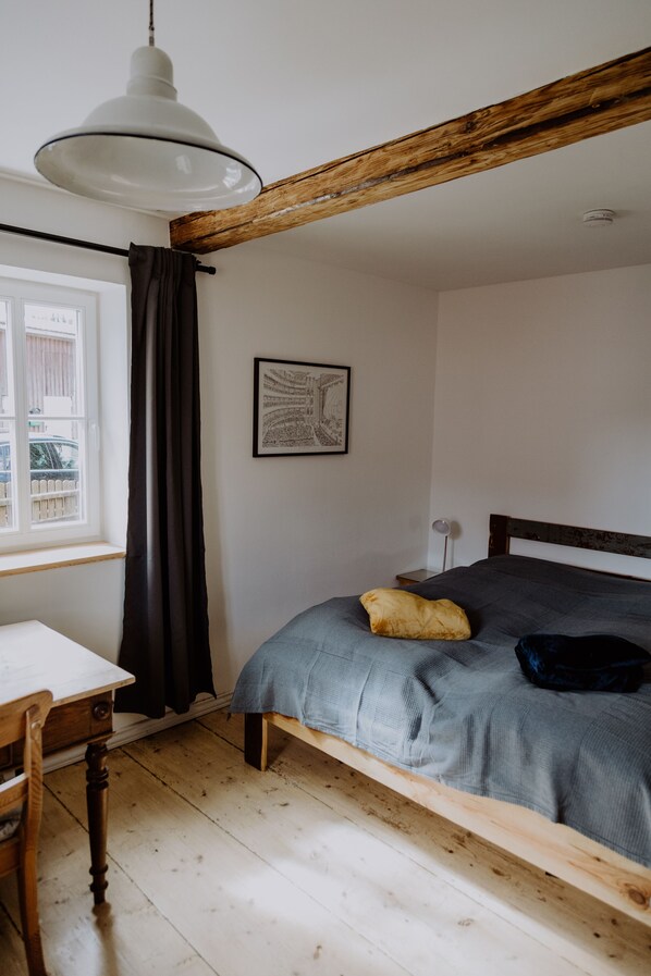 A bedroom with a wooden ceiling, a bed with a grey blanket, a bedside table, a chair, a window with curtains, and a framed picture on the wall. - Lovingly Renovated Farmhouse in Rhön/Kaltenwestheim (Kaltennordheim)