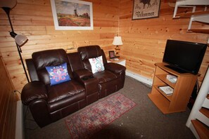 L'intérieur d'un chalet accueillant, avec un canapé en cuir marron, un téléviseur posé sur un meuble en bois, une lampe et un panneau d'accueil encadré.