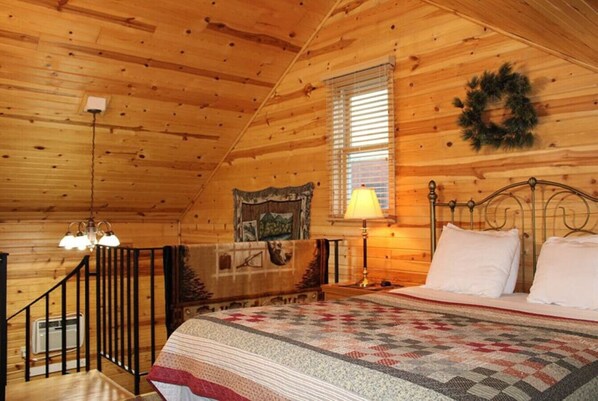 A cozy bedroom with a wooden ceiling, a bed with a quilt, a nightstand, a lamp, and a window with blinds.