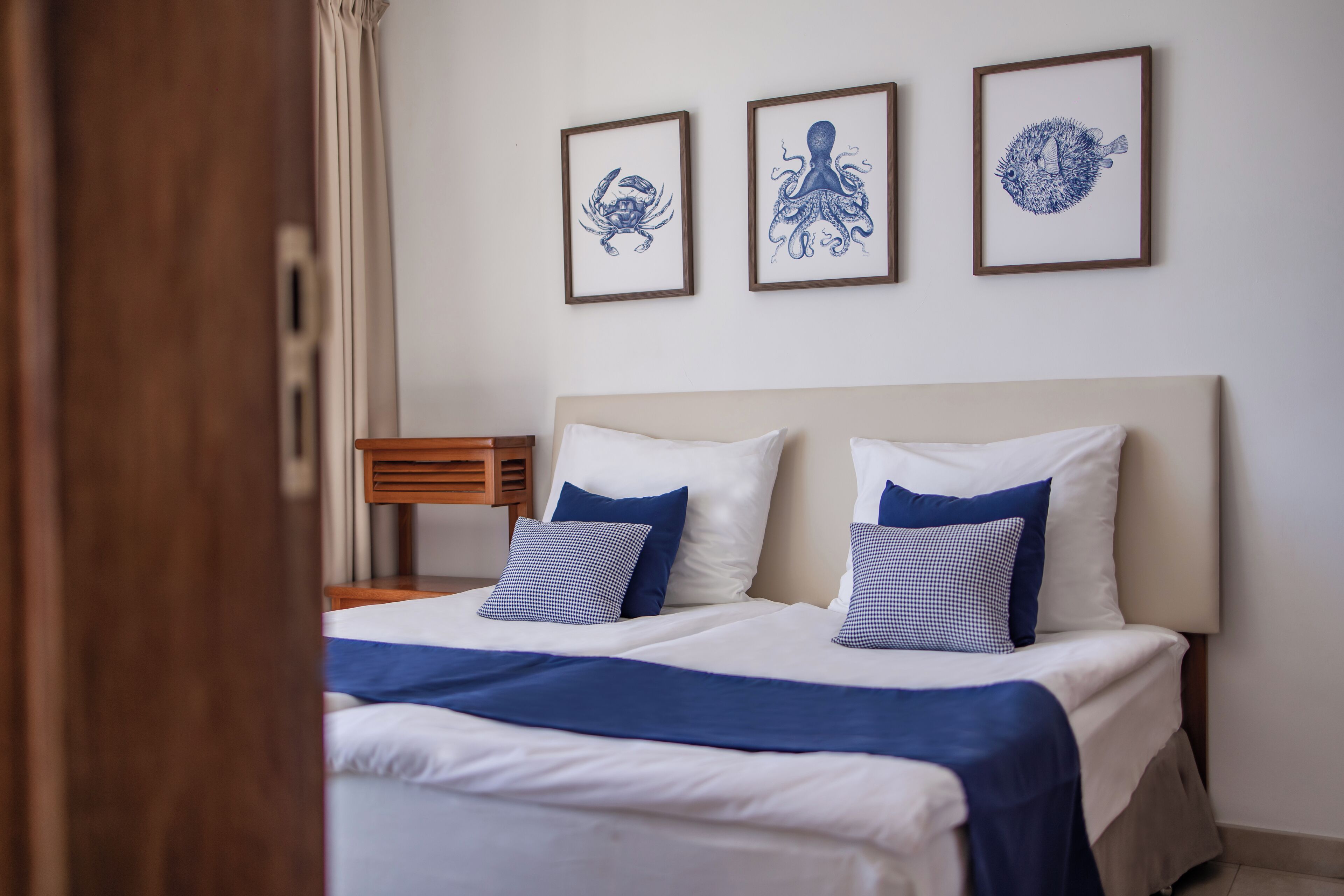 A neatly made bed with white and blue bedding, a wooden nightstand, and three framed artworks on the wall.