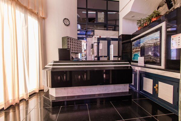 A reception area with a black and white checkered floor, a clock on the wall, and a large window with blinds. - Servatur JB - Adults Only (Mogan)