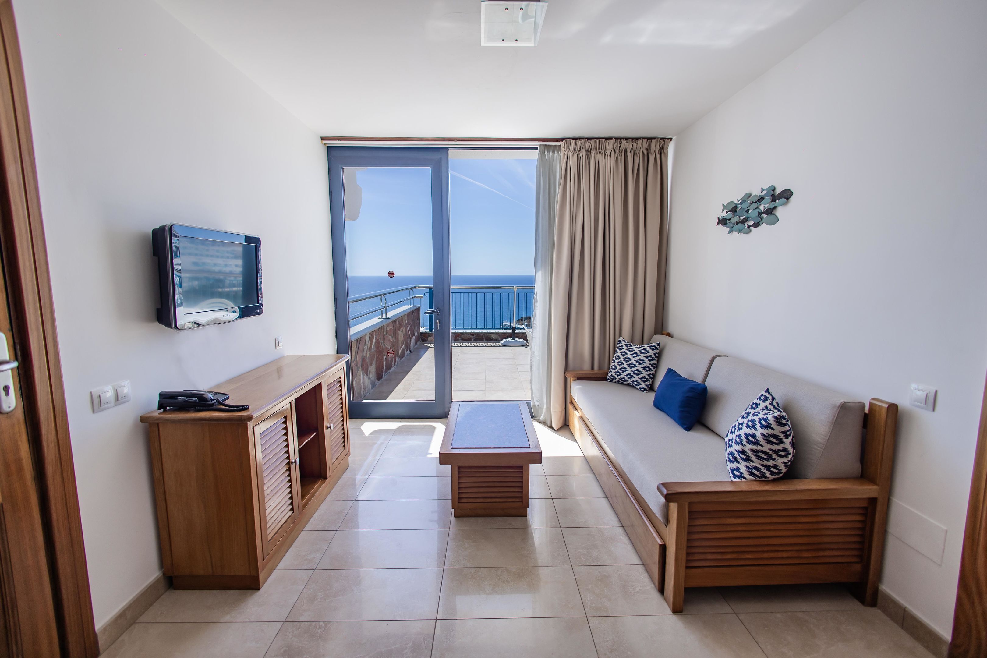 A room with a wooden bench, a sofa with patterned cushions, a television mounted on the wall, and a view of the sea through a glass door.