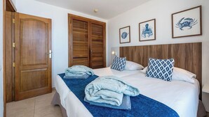 A hotel room with a wooden door, a bed with blue bedding, two towels on the bed, and three framed pictures on the wall. - Servatur JB - Adults Only (Mogan)