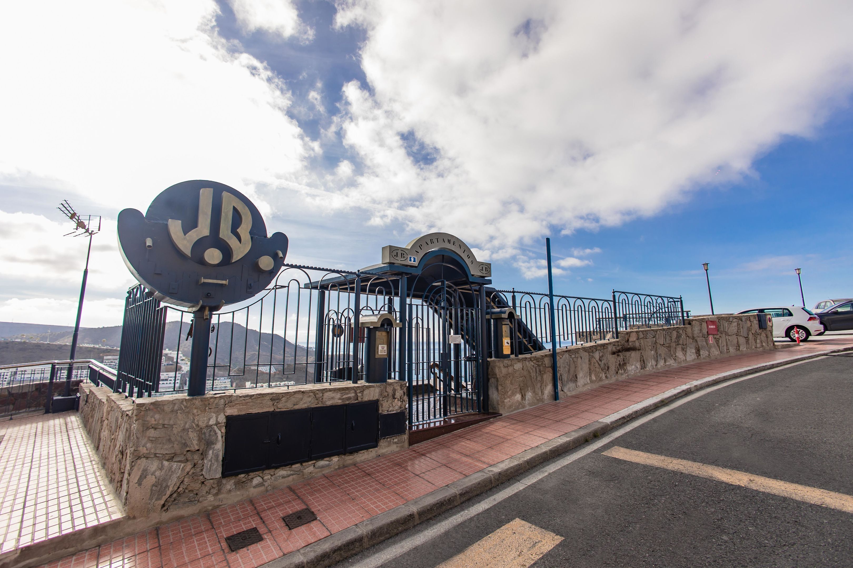 Un paseo marítimo con barandilla de metal, muro de piedra y un cartel con un emblema circular.