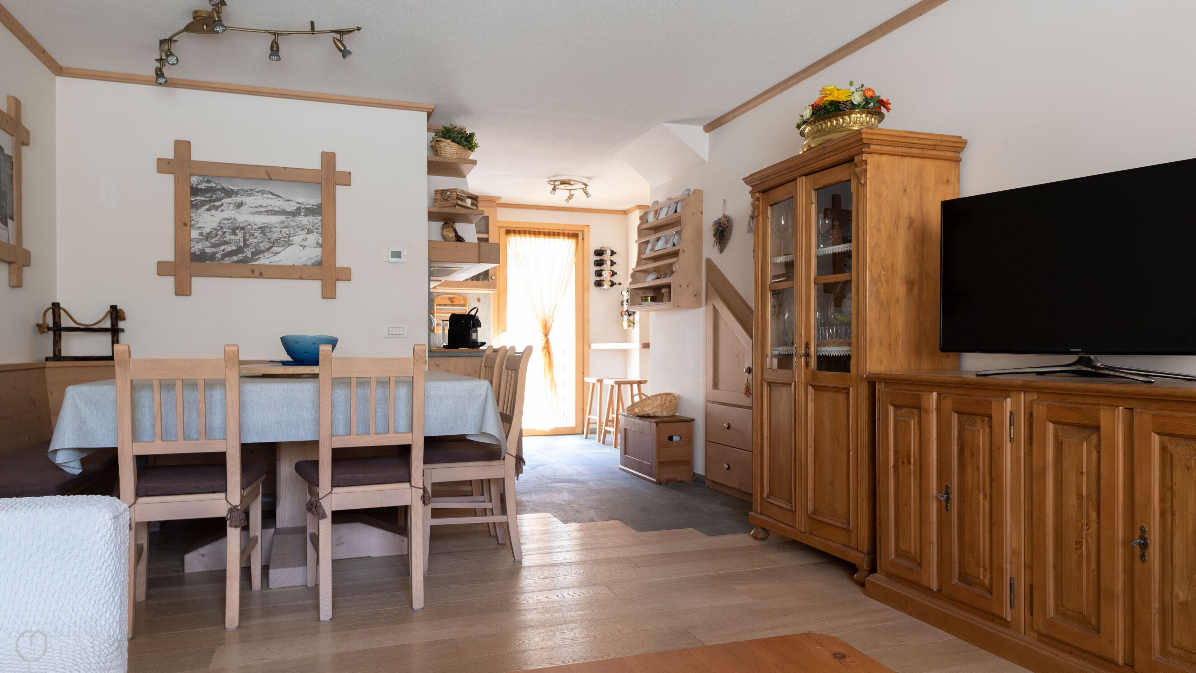 Zona pranzo con tavolo e sedie in legno, televisore su un mobile in legno e cucina sullo sfondo.