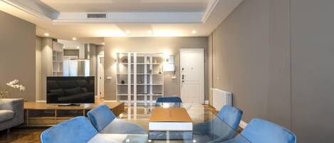 A modern living room with a glass-top coffee table, blue upholstered chairs, a flat-screen TV, and a bookshelf.