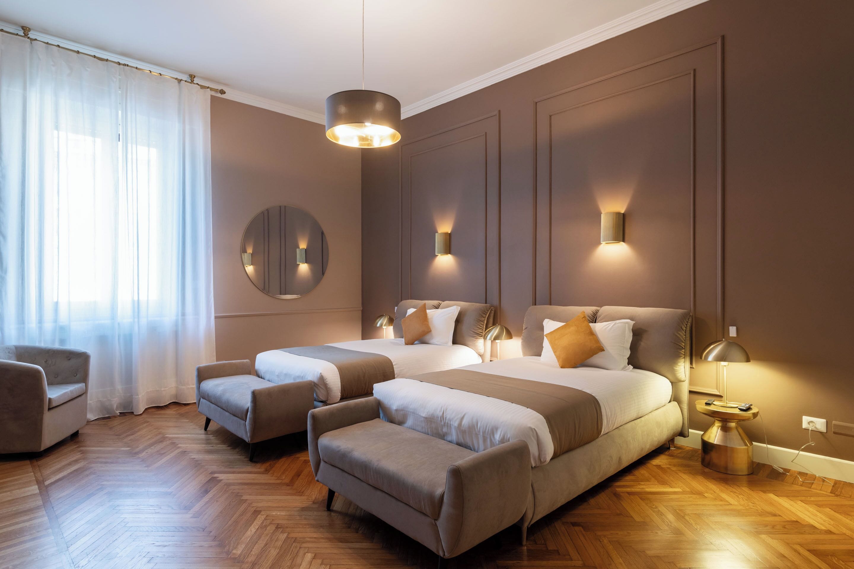 A hotel room with two beds, a round mirror, wall sconces, a side table, and a wooden floor.