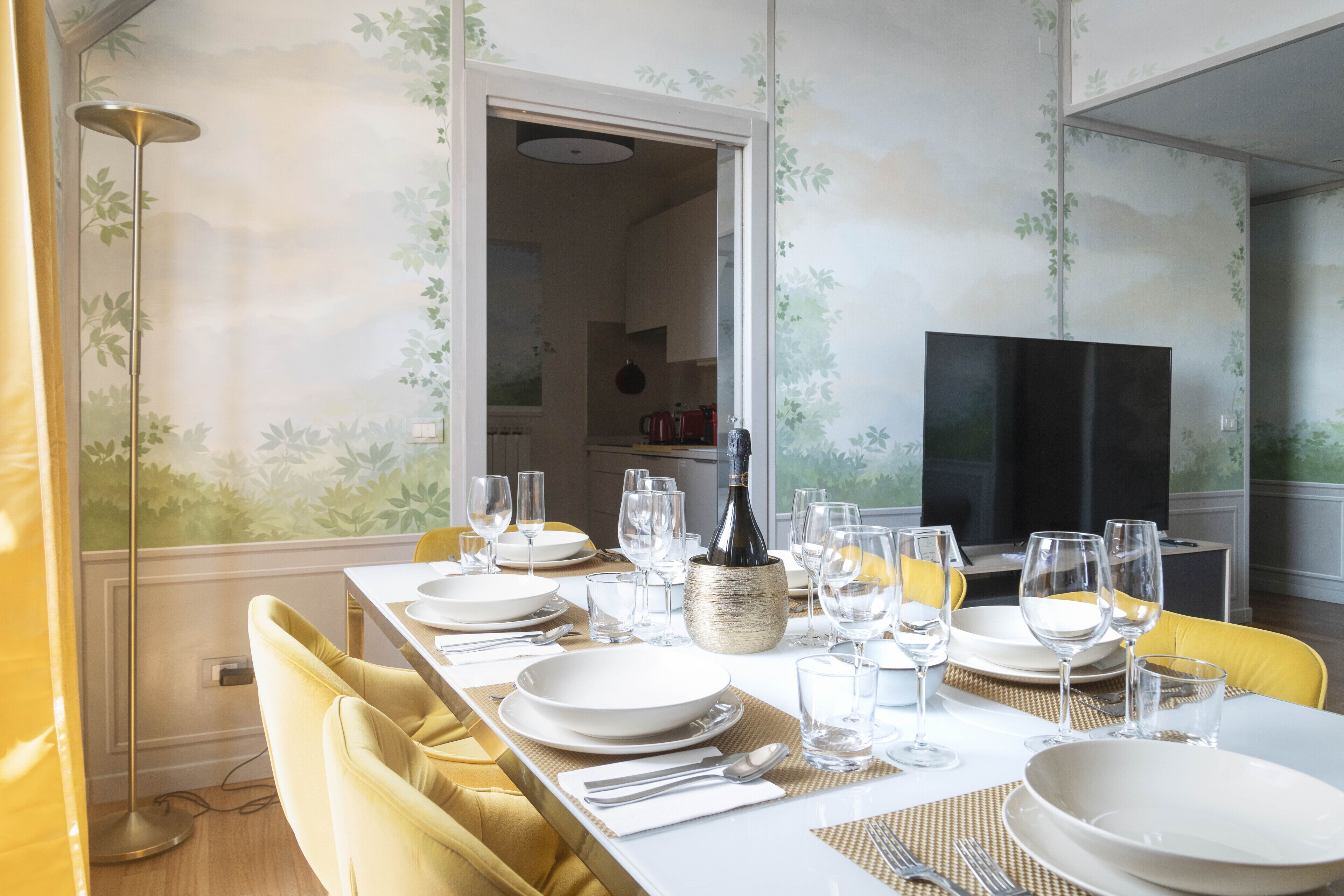 A dining room with a long table set for six, a TV in the background, and a kitchen visible through an open doorway.
