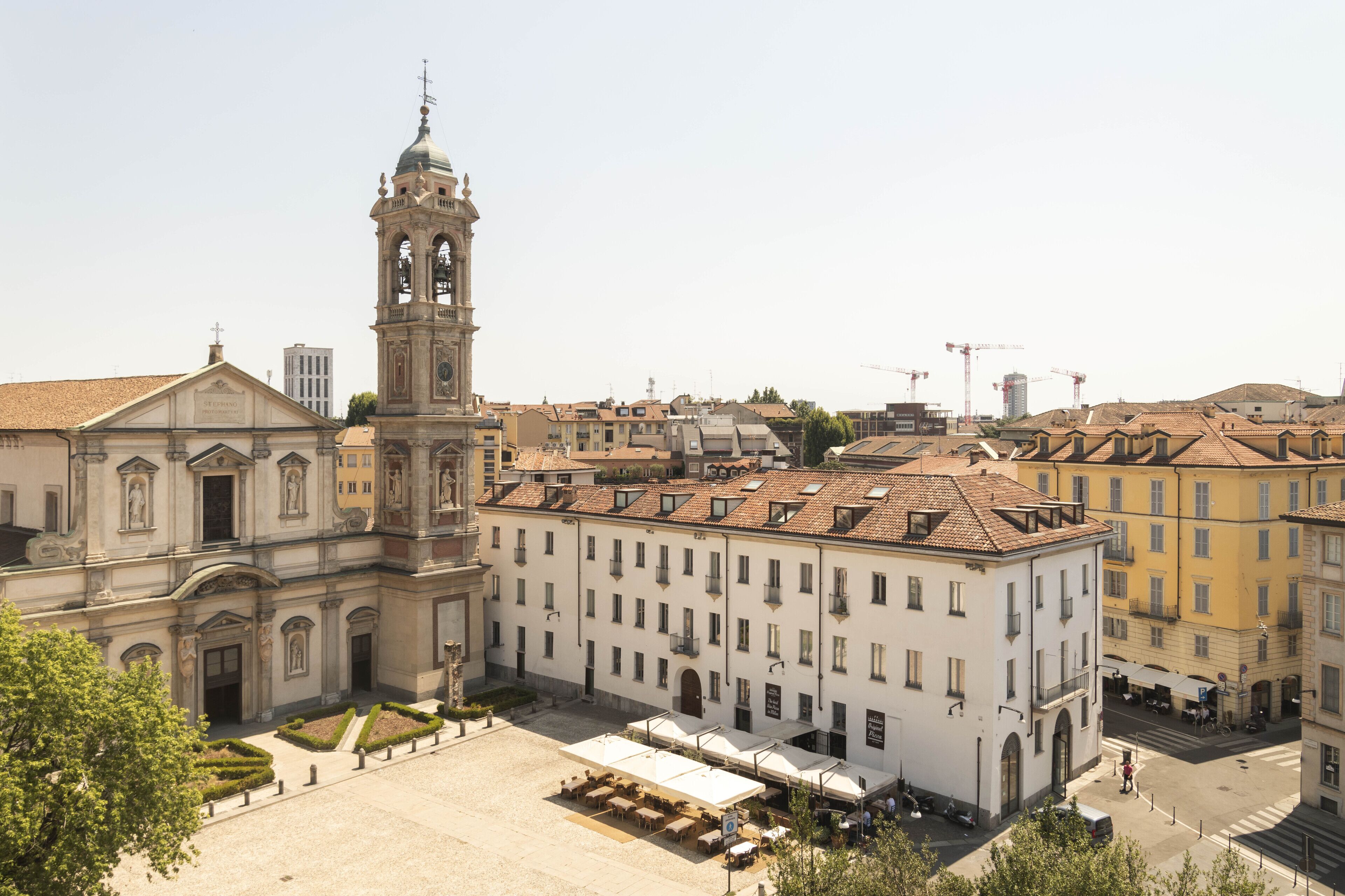 A historic building with a bell tower, a courtyard with outdoor seating, and a street with construction cranes in the background.