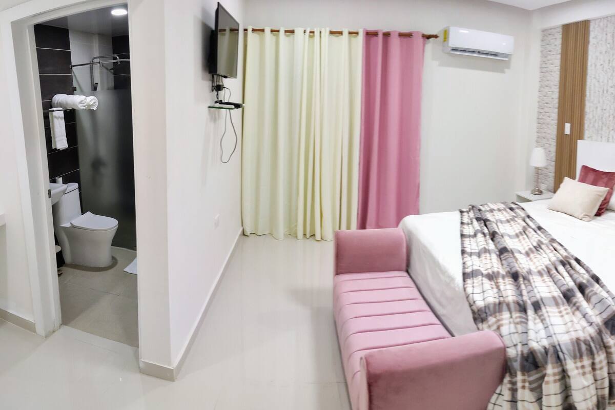 Une chambre d’hôtel moderne équipée d’un grand lit, d’une table de chevet, d’une petite commode, d’un bureau, d’une chaise, d’un espace douche et de toilettes.