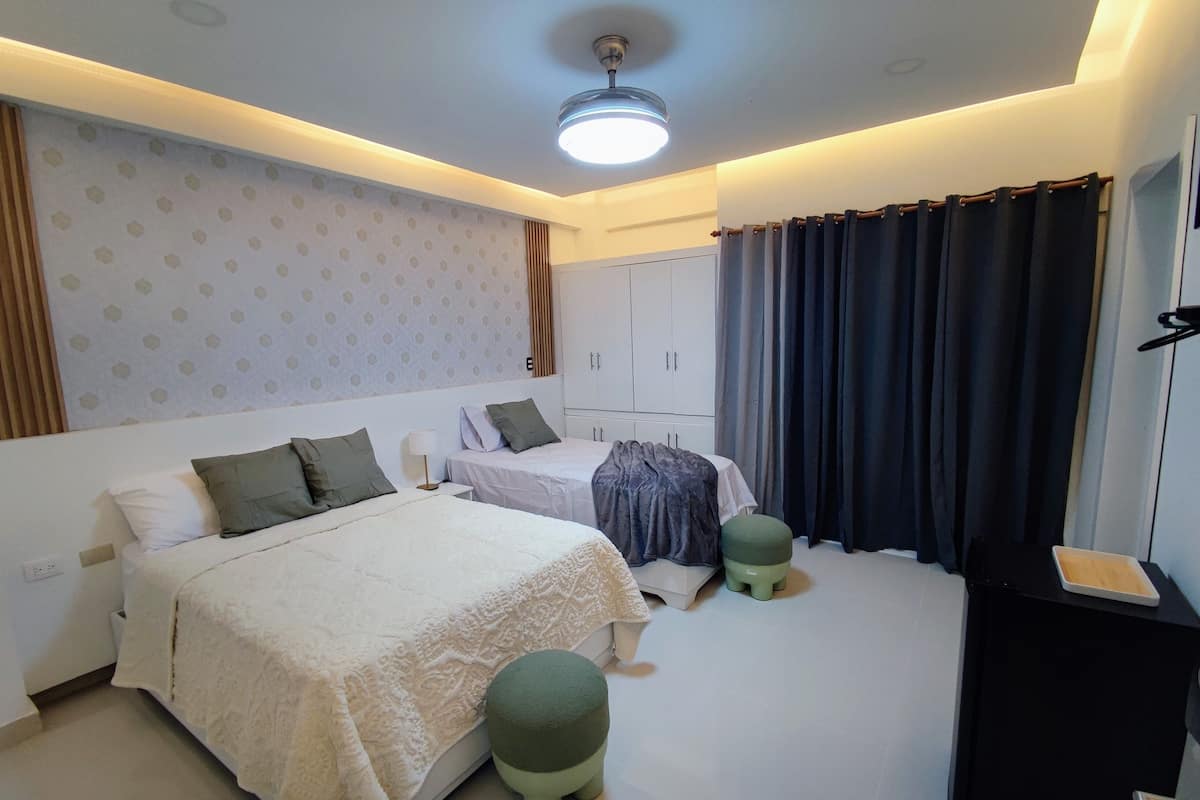 Une chambre d’hôtel avec deux lits, un bureau, une chaise, une armoire et une grande fenêtre avec des rideaux.