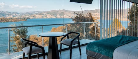 Un dormitorio con una cama grande, una mesita, dos sillas y un televisor. La habitación tiene un balcón con vistas a un lago y montañas.