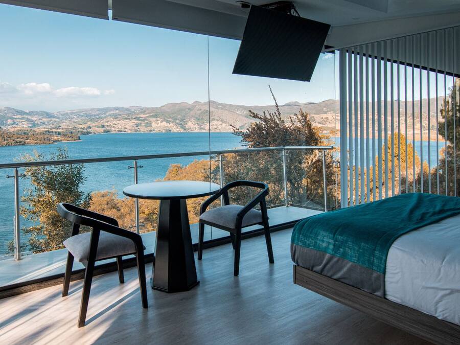 A bedroom with a large bed, a small table, two chairs, and a TV. The room has a balcony with a view of a lake and mountains.