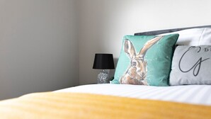 A neatly made bed with a yellow blanket, a black lamp, and two pillows with a rabbit design and a partial text on one pillow.