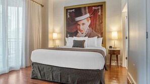 A neatly made bed with white linens and a gray skirted bedspread, flanked by two bedside tables with lamps, in a room with a large framed portrait on the wall. - Innovative Décor in heart of Theater District! (New Orleans)