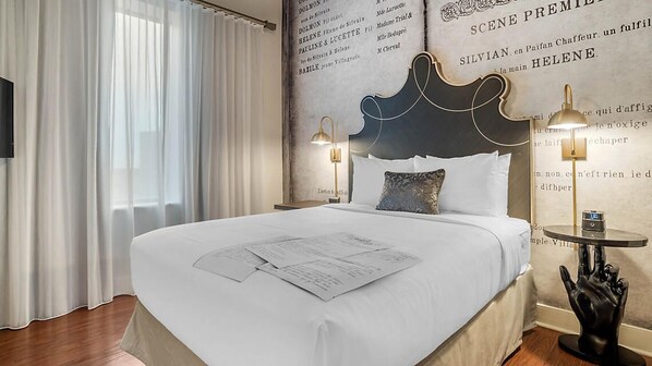 A hotel room with a large bed, a headboard with a decorative design, two bedside lamps, and a wall with text and a hand sculpture. - Innovative Décor in heart of Theater District! (New Orleans)