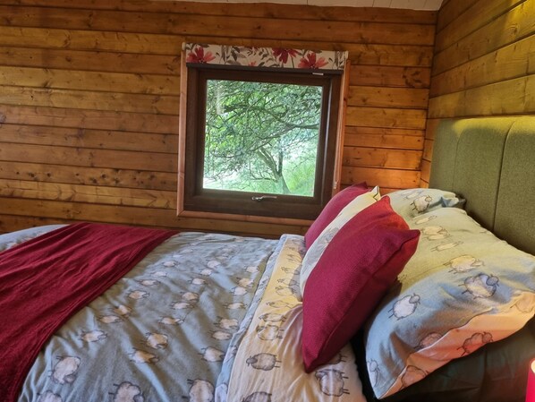 Une chambre dans un chalet en bois, avec un lit, une fenêtre donnant sur la forêt et une tête de lit recouverte d’un tissu vert.
