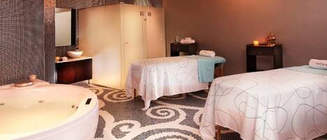 Habitación de hotel con una bañera grande, dos camas con cabeceros iguales, un tocador con espejo y una mesita de noche con artículos de aseo.