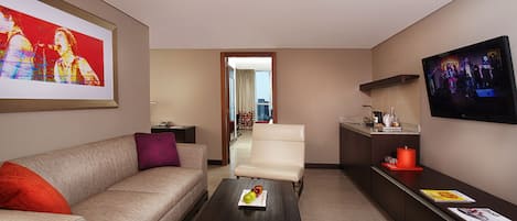 A modern hotel room with a sofa, armchair, coffee table, TV, and a city view through the door.