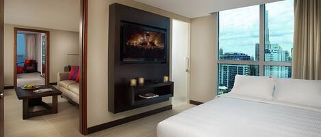 Habitación de hotel con una cama grande, un televisor montado en la pared, un rincón de descanso con un sofá y una mesita de centro, y vistas a la ciudad a través de amplias ventanas.