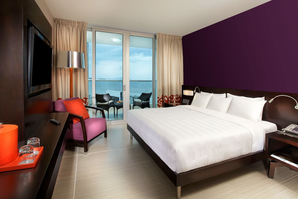 A hotel room with a large bed, a desk with a computer, a TV, and a balcony with chairs.