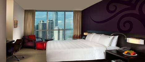 Habitación de hotel con una cama grande, un escritorio, una silla, una mesita de noche y vistas del horizonte de la ciudad.