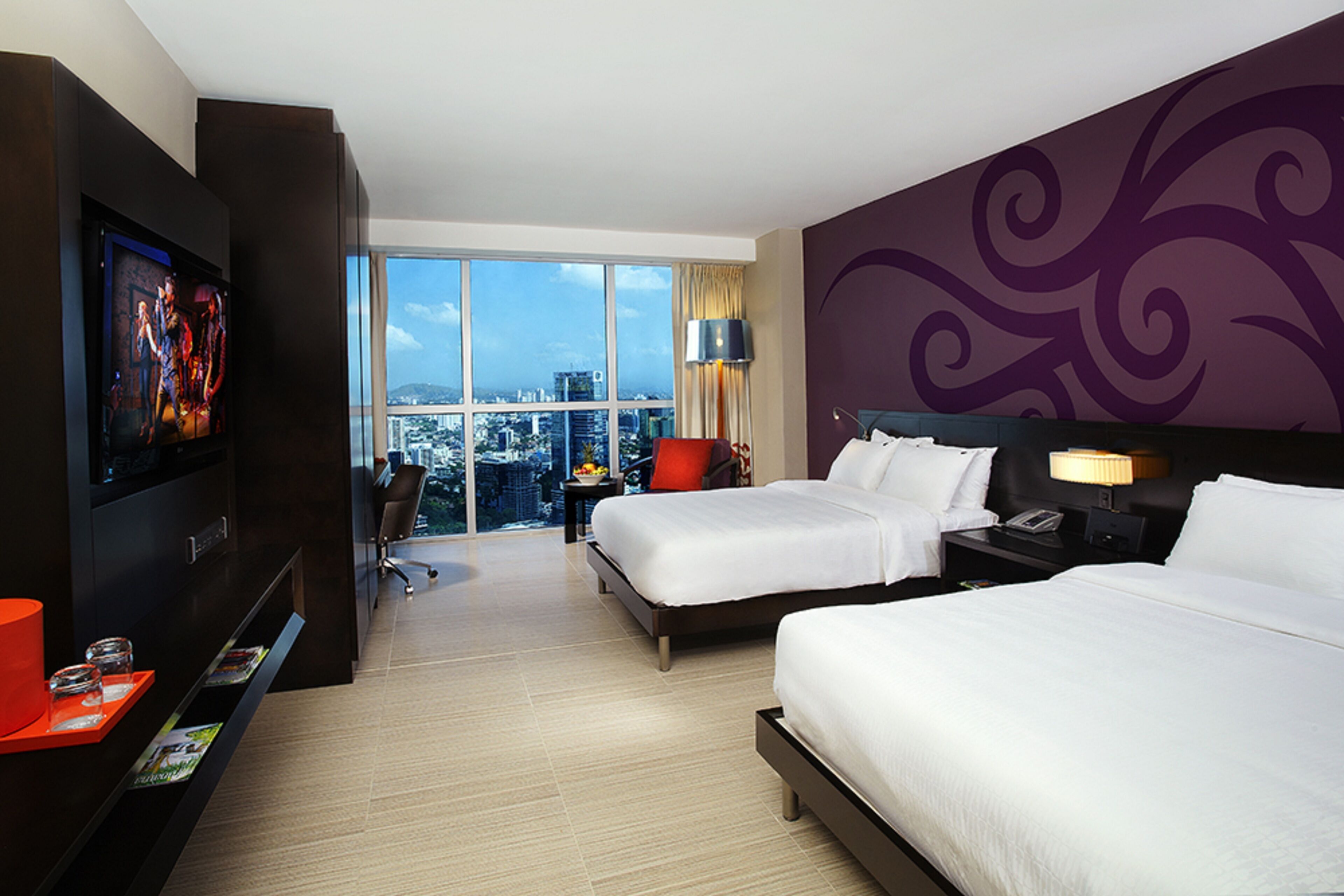 Habitación de hotel con dos camas, televisión, escritorio y un amplio ventanal con vista a la ciudad.