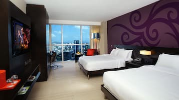 Habitación de hotel con dos camas, televisión, escritorio y un amplio ventanal con vista a la ciudad.