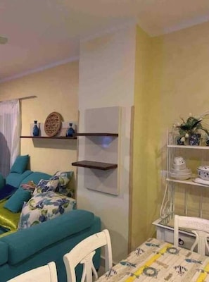 Un salon chaleureux avec un canapé de couleur turquoise, une table blanche recouverte d’une nappe à motifs et des étagères ornées d’objets décoratifs.