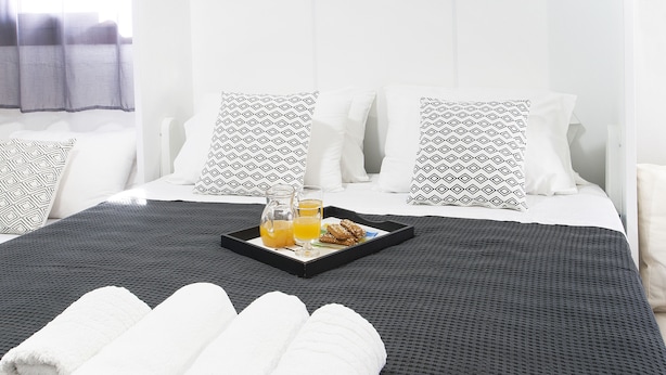 Ein ordentlich bezogenes Bett mit einer schwarz-weiß gemusterten Tagesdecke, weißen Kissen und aufgerollten weißen Handtüchern. Ein Tablett mit Orangensaft und Gebäck steht auf dem Bett.