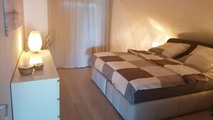 Une chambre à coucher comprenant un lit, une table de chevet avec une lampe, une fenêtre avec des rideaux et une étagère fixée au mur.
