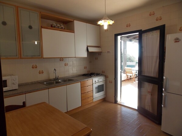 Een kleine keuken met witte kastjes, een spoelbak, een fornuis en een magnetron. Er staat een houten tafel met een stoel, en een deur naar een buitenruimte.