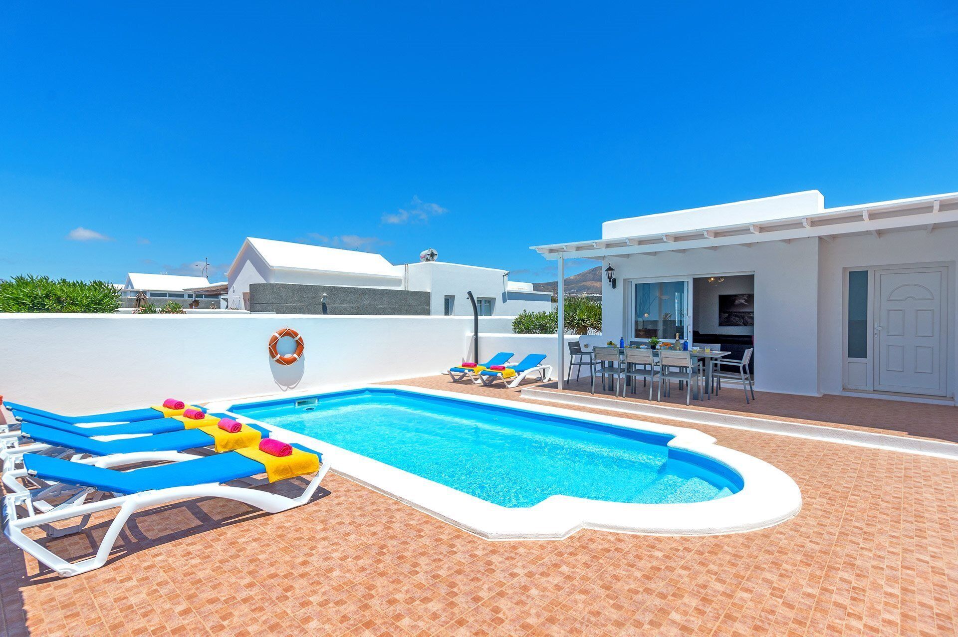 A pool area with lounge chairs and a table, surrounded by white buildings and a clear blue sky.