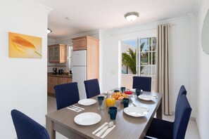 A dining area with a table set for four, a kitchen in the background, and a window with a view of palm trees.