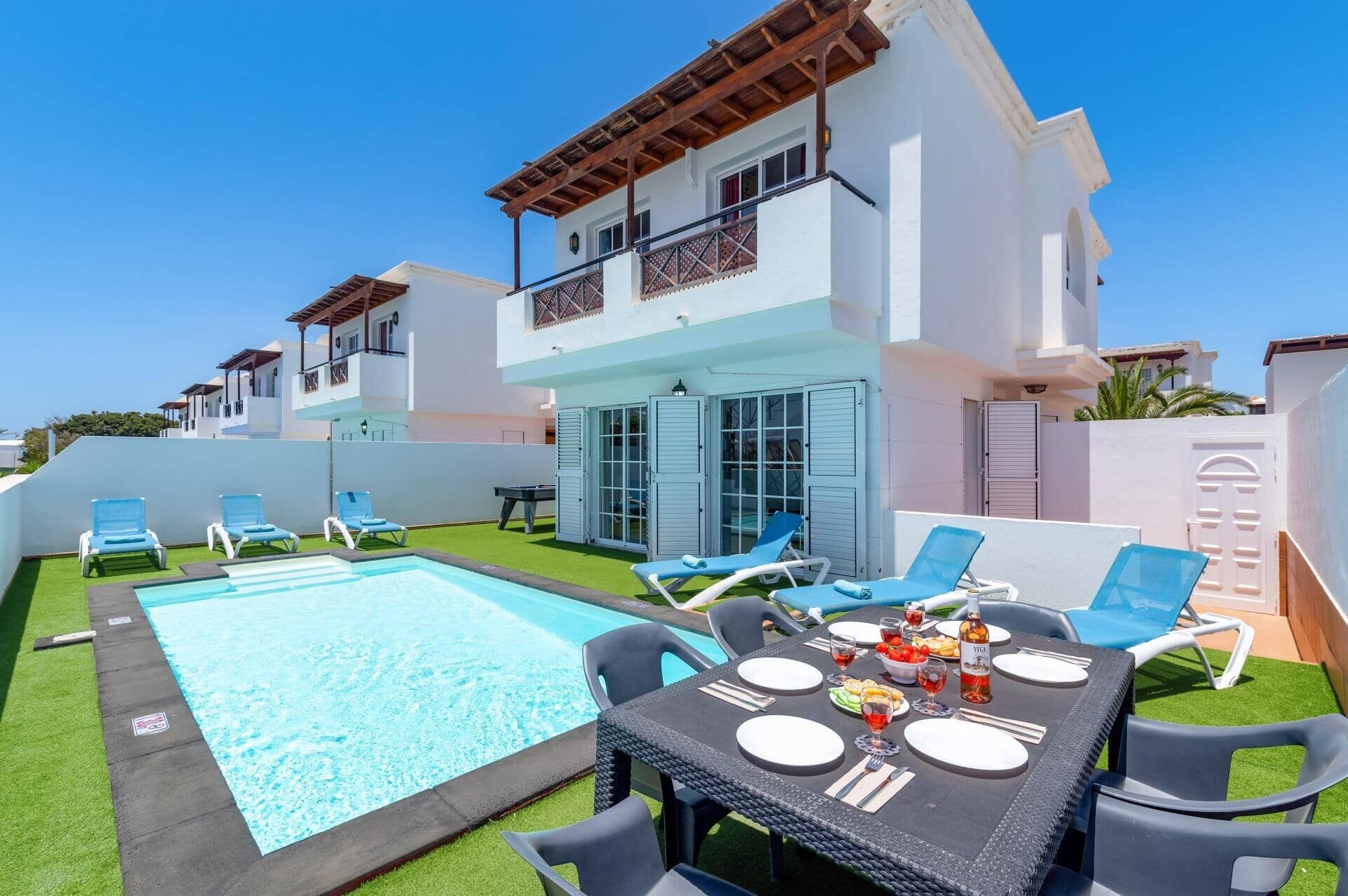A poolside area with a dining table set for two, a pool, and a white building with balconies.