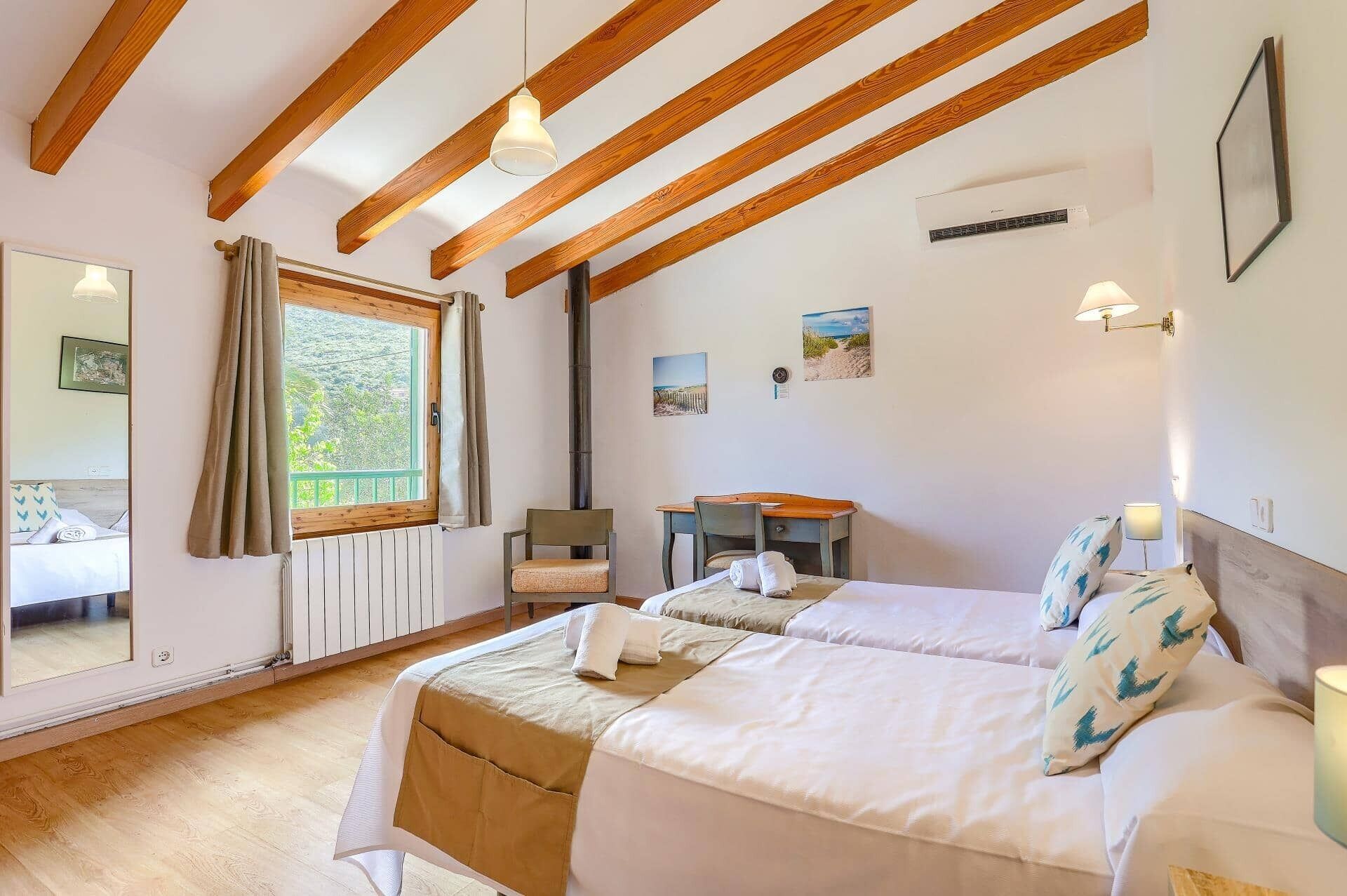A bedroom with two beds, wooden beams, a window with curtains, a desk, and wall-mounted lights.
